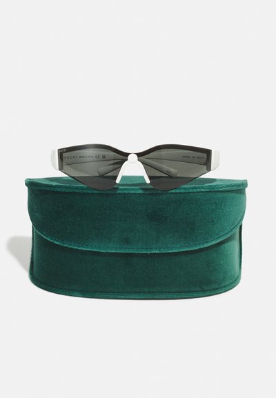 Gucci-solbriller med svart linse, unik bølget form og hvite detaljer, plassert på et grønt fløyelsetui med avrundede kanter.