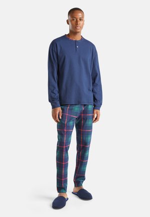 Mann trägt ein marineblaues langärmeliges Henley-Shirt, grün-rot karierte Pyjamahose und marineblaue Hausschuhe, steht vor weißem Hintergrund.