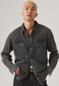 Camisa de mezclilla gris oscuro con diseño de botón, dos bolsillos en el pecho y detalles de costura más clara en contraste.