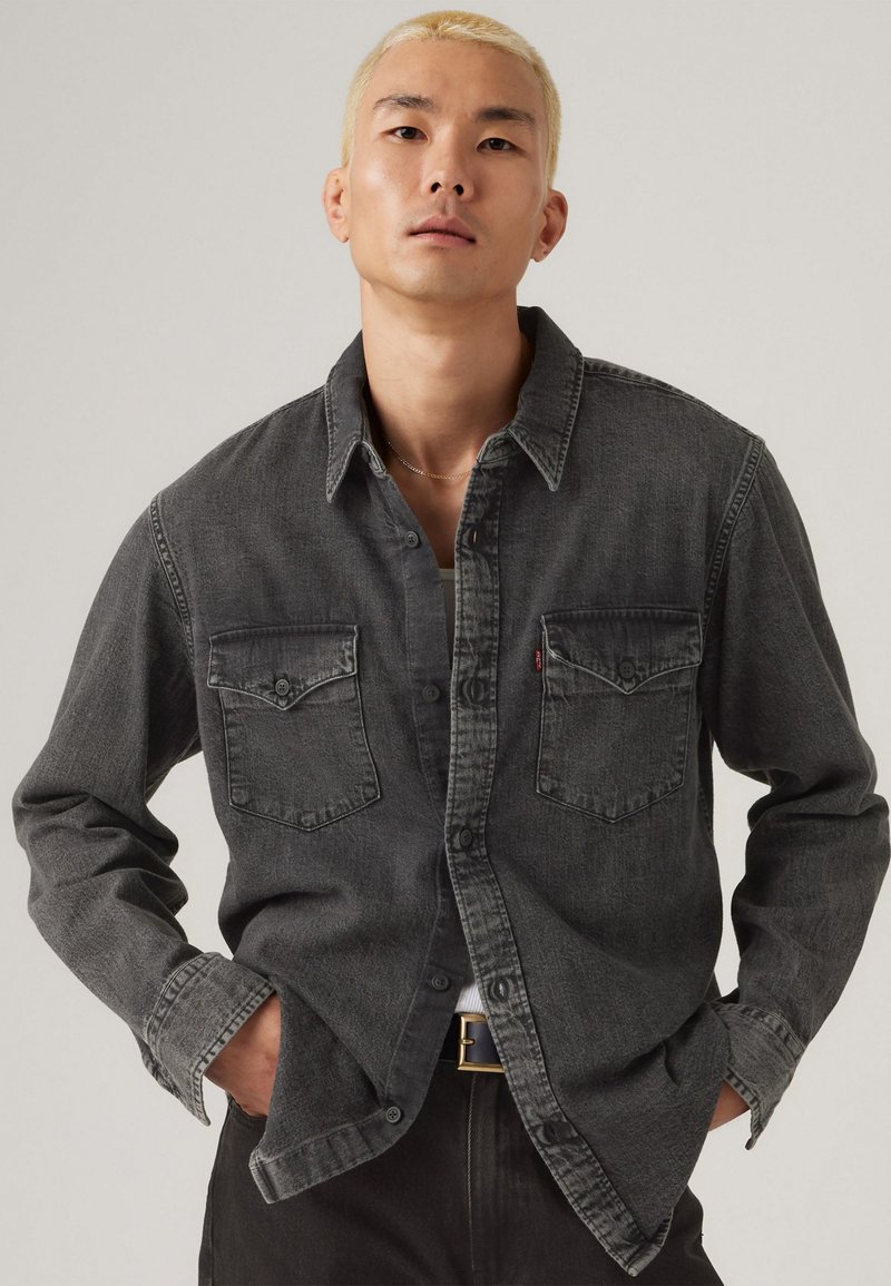 Camisa de mezclilla gris oscuro con diseño de botón, dos bolsillos en el pecho y detalles de costura más clara en contraste.