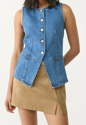 Mouwloos blauw denim gilet met knopen en zakken, gecombineerd met een lichtbruine suède minirok met een kleine zijsplit.