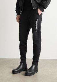 Crne sportske hlače od mekog materijala, s bijelim "Calvin Klein" logotipom sa strane, u kombinaciji s elegantnim crnim čizmama do gležnja.