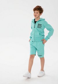 Turquoise sweatshirt met capuchon en voorzakken, voorzien van zwarte tekst; bijpassende shorts in een relaxte pasvorm; witte sneakers.
