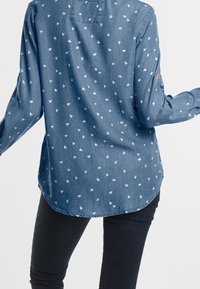 Jeanskjorta, ljusblå med vita fågelmönster, långa ärmar, rundad nederkant, subtil bröstficka, uppvikta ärmslut.