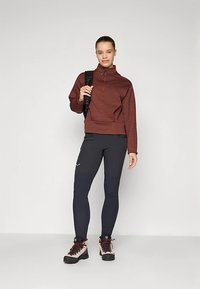 Brūns pulovers ar pusritenti un rāvējslēdzēju, melnas elastīgas bikses un pārgājienu zābaki. Modelis tur melnu mugursomu, stāvot uz neitrālas krāsas fona.