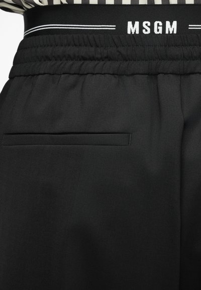 Schwarze Hosen mit strukturierter Oberfläche, elastischem Bund und einem weißen Logo. Beinhaltet eine flache Tasche auf der Rückseite.