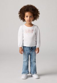 Camicia bianca a maniche lunghe con un logo Levi's colorato, abbinata a jeans svasati blu chiaro e sneakers bianche. Outfit semplice e casual.