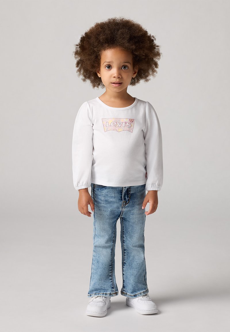 Camicia bianca a maniche lunghe con un logo Levi's colorato, abbinata a jeans svasati blu chiaro e sneakers bianche. Outfit semplice e casual.