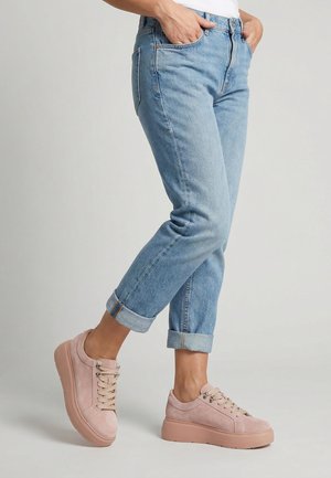 Person iført lyseblå jeans med opsmøgede ben og pastelfarvede lyserøde plateau-sneakers, stående med hænderne i lommerne.