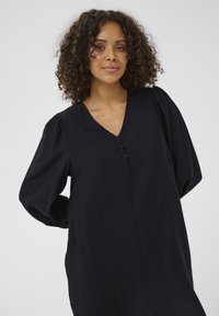Robe noire à manches longues avec un col en V et une patte de boutonnage. Tissu texturé et manches bouffantes pour une silhouette détendue.