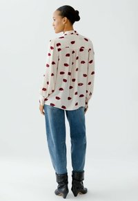 Leichtes, creamfarbenes Blusenshirt mit roten, lippenförmigen Mustern, langen Ärmeln und einem geknöpften Rücken. Kombiniert mit lockeren blauen Jeans und schwarzen Stiefeln.