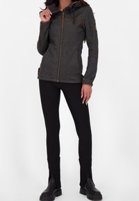 Šedá zipová bunda s kapucí, hnědými akcenty a prošíváním; spárována s černými upnutými kalhotami se štěrbinkami na lemu a botami do výšky kotníku.