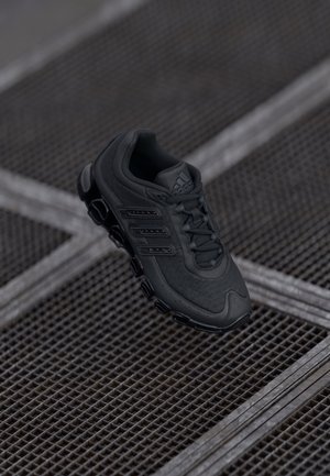 Chaussure de sport noire Adidas avec semelle texturée suspendue au-dessus d'un sol en grille métallique dans un environnement peu éclairé.