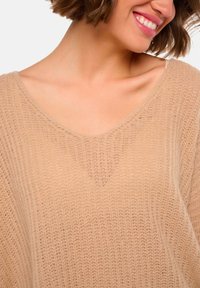 Pull en tricot beige avec un large décolleté en V, doté d'une texture douce et légère et d'une coupe ample. Motif de couture vertical visible.