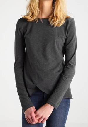 Frau mit blonden Haaren, die ein dunkelgraues Langarmshirt und blaue Jeans trägt, steht mit vor dem Körper verschränkten Händen.