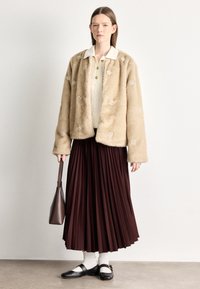 Casaco bege de pelo falso com aplicações florais, por cima de um top creme, combinado com uma saia plissada bordô e sapatos pretos com tiras.