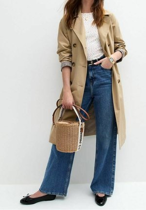 Frau trägt beigen Trenchcoat, weiß gemustertes Top, blaue weite Jeans, schwarze Ballerinas, hält eine geflochtene Eimertasche mit Lederriemen.