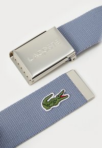 Lacoste blå vevet belte med en sølvfarget metallspenne. Har et brodert grønt krokodille-logo. Teksturert stoff, slank design.