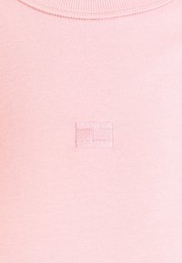 Camiseta de algodón rosa claro con un escote acanalado y una pequeña etiqueta de logo cuadrada cosida en el lado izquierdo. Textura suave, diseño casual.