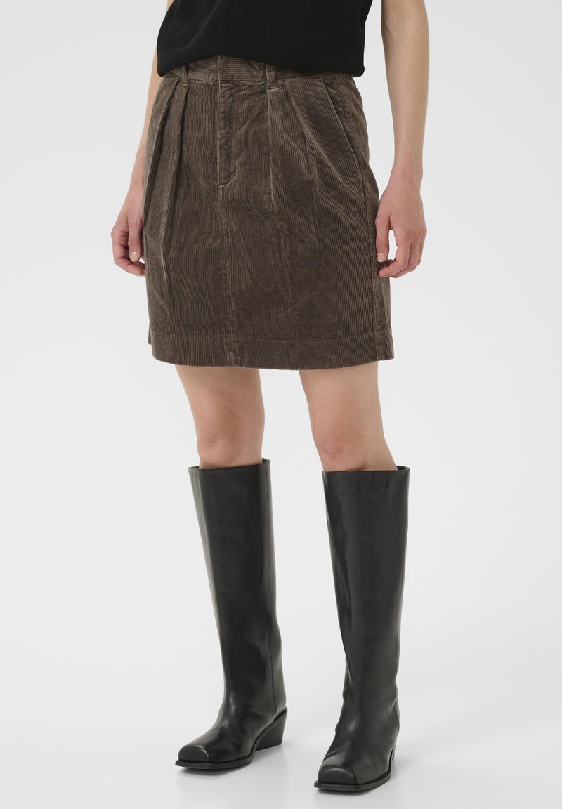 Jupe en velours côtelé marron avec des plis devant et une coupe droite, associée à des bottes en cuir noires montantes jusqu'aux genoux avec un talon compensé.
