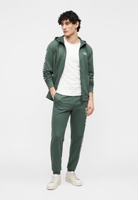 Groene hoodie met rits en bijpassende joggers gemaakt van zachte stof. Wit T-shirt eronder. Sneakers. De hoodie heeft een logo op de borst.