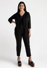Zwarte opengehaakte cardigan over een zwarte top, gecombineerd met zwarte fitted broek en zwarte platform sandalen met houten hakken.