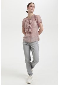 Giovane donna in una blusa di fiori con volant, jeans grigi attillati e sneakers bianche, in piedi con le mani dietro la schiena su sfondo bianco.