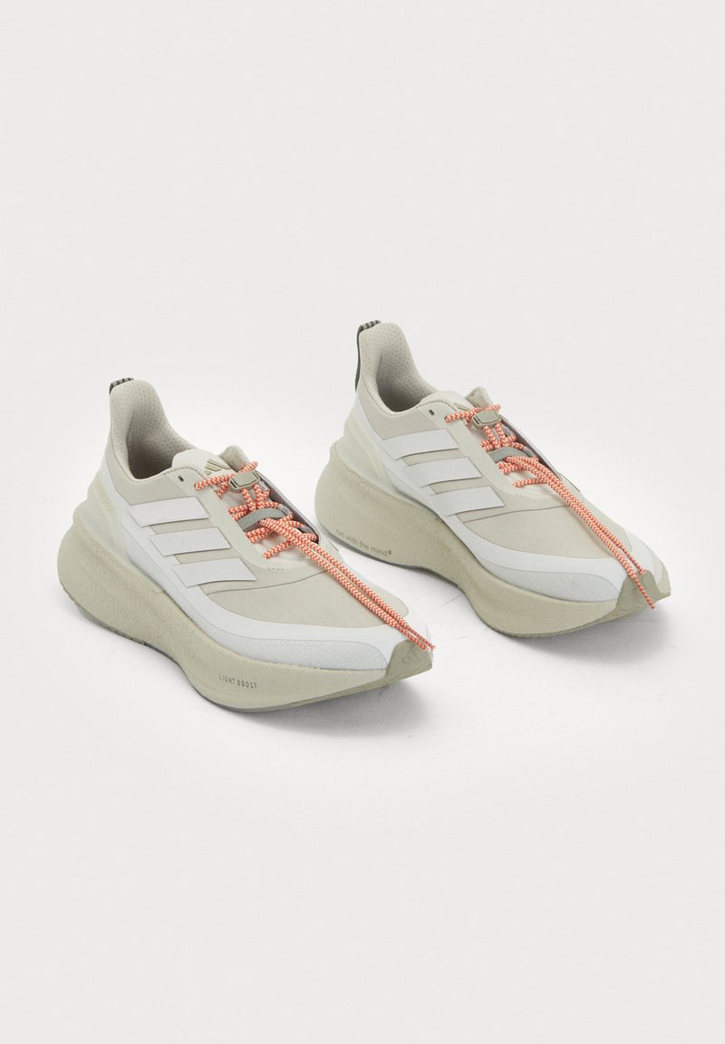 Coppia di scarpe sportive color avorio con suole spesse, tre strisce laterali bianche e lacci arancioni e bianchi su uno sfondo chiaro uniforme.