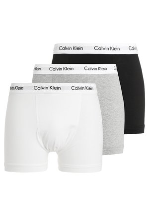 Jeune homme portant un t-shirt blanc Calvin Klein, un boxer gris et des chaussettes blanches, soulève son t-shirt pour révéler le logo à la taille.