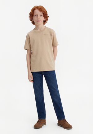 Παιδί με κόκκινα μαλλιά, που στέκεται φορώντας μπεζ κοντομάνικο πουκάμισο, μπλε τζιν και καφέ μπότες με κορδόνια, σε λευκό φόντο.