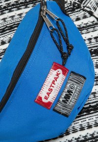 Borsa in tessuto blu con due scomparti con zip, dotata di toppe per logo rosse e nere, materiale testurizzato e dettagli in metallo neri.