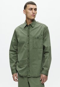 Camicia di cotone verde oliva con bottoni, taschino sul petto e colletto a punta. Caratterizzata da una vestibilità rilassata e dettagli cuciti sui polsini.