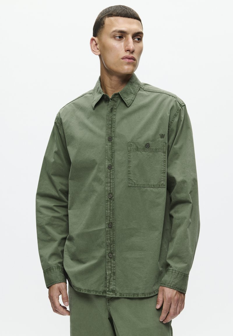 Camisa de algodão verde-oliva com botões, um bolso no peito e colarinho pontudo. Apresenta um corte relaxado e costura decorativa nos punhos.