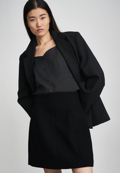 Blazer noir ajusté porté sur un haut assorti et une jupe taille haute. Tissu texturé, design structuré et détails de boutons.