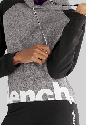 Personne portant un sweat à capuche Bench gris et noir et un pantalon noir, tenant le cordon du sweat à capuche d'une main.