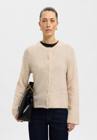 Light beige cardigan made of soft material, featuring a round neckline, five button closure, and long sleeves. Paired with a black bag.