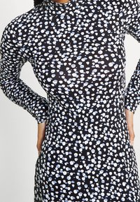 Svart klänning i tyg med lång ärm, med ett mönster av blå och vita prickar. Hög halsringning, figurnära silhuett, mjuk textur.