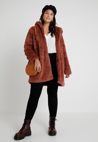 Rudas dirbtinio kailio paltas su kapišonu, dėvimas virš baltos maikos ir juodų kelnių. Raštuota medžiaga, ilgomis rankovėmis ir priekiniais atidarymais. Ruda per petį nešama kuprinė.