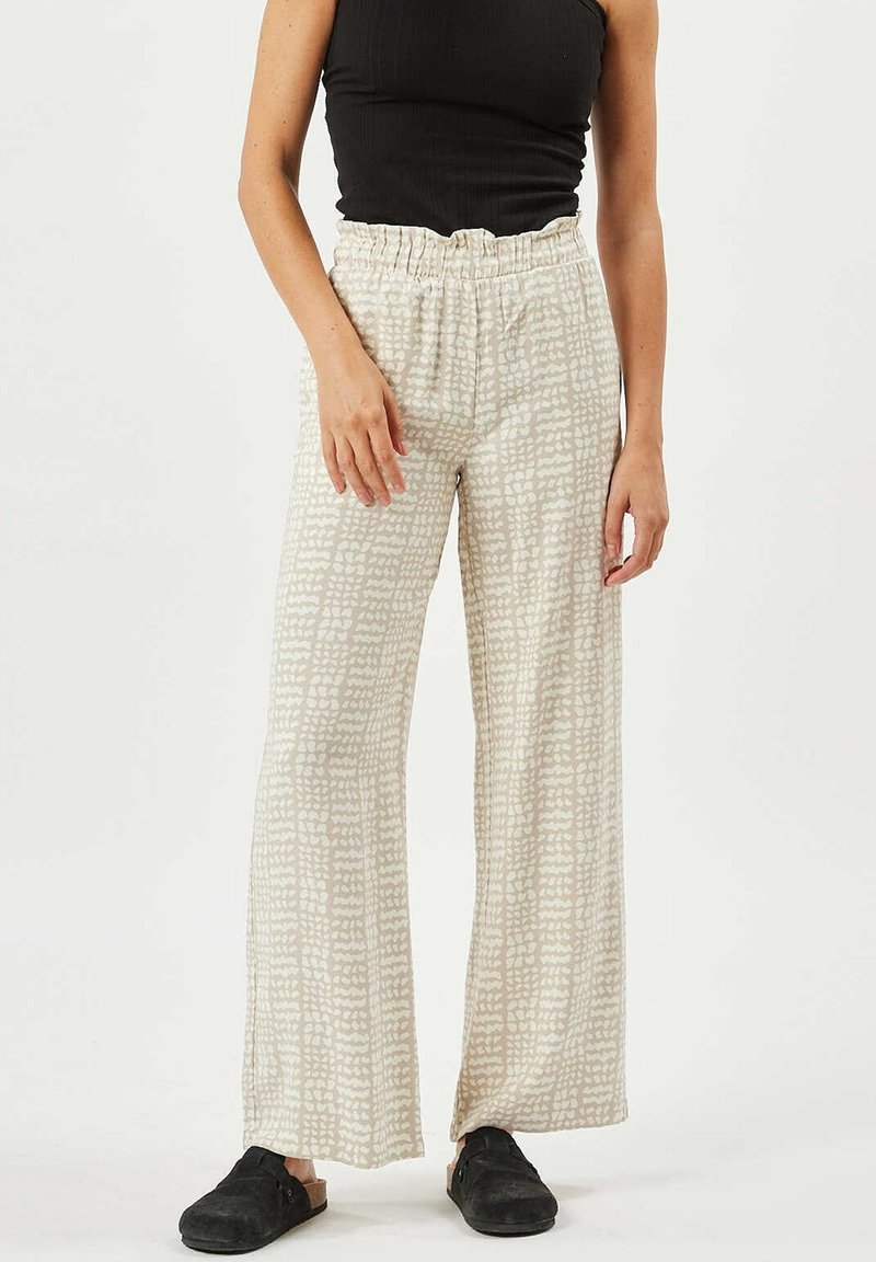 Femme portant un pantalon large beige à motifs, un haut noir sans manches et des chaussures noires à enfiler sur un fond clair uni.