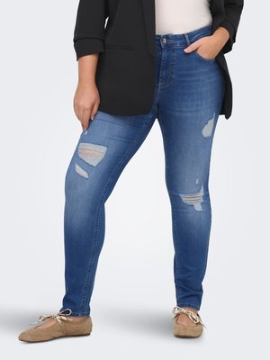 Γυναίκα με σκισμένο μπλε skinny τζιν, ταμπά μπαλέτα με κορδέλες, μαύρο σακάκι και λευκό τοπ, στέκεται μπροστά σε απλό φόντο.