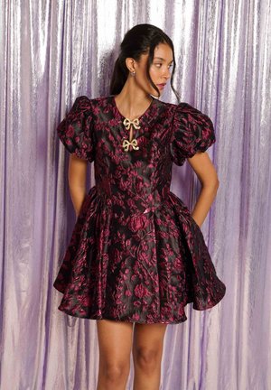 Femme en robe fleurie noire et rouge foncé avec des manches bouffantes et deux broches nœuds dorés, debout devant un rideau lavande brillant.