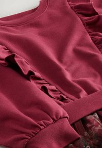 Burgundská mikina s volánkovými akcenty na rukávech, elastickými manžetami a texturovaným materiálem; doplněna květinovým tylovým lemem na spodní části.