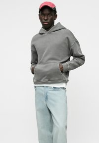 Γκρι φούτερ με κουκούλα, μπροστινές τσέπες και ρεβέρ στους καρπούς, σε συνδυασμό με ανοιχτό μπλε τζιν. Το άτομο φορά ένα ροζ καπέλο μπέιζμπολ.