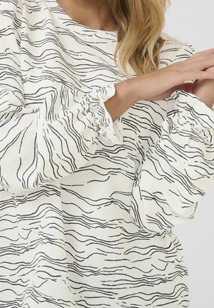 Vrouw die de mouw van een witte blouse met zwarte, onregelmatige, golvende lijnpatronen en geplooide elastische manchetten aanpast.