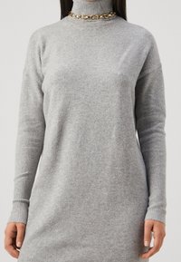 Femme portant une robe-sweater à manches longues gris clair avec des poignets côtelés et une chaîne en or, se tenant devant un fond uni.
