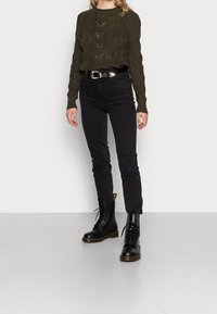 Pull en maille vert olive avec motif texturé, associé à un jean noir taille haute et des bottes noires à lacets avec semelles épaisses.