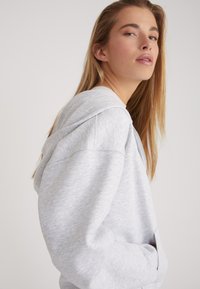 Lichtgrijze hoodie van zacht materiaal, met een grote capuchon, een voorzak en ribgebreide mouwen, ontworpen met een ontspannen pasvorm.