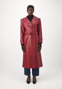 Lang rød læder trench coat med dobbeltradet design, bælte i taljen, skulderklaffer og glat tekstur. Båret over en mørk top og jeans.