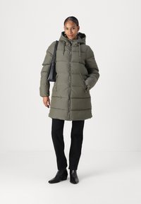 Manteau rembourré vert olive avec capuche, motif matelassé, fermeture éclair à l'avant, poches latérales ; associé à un pantalon noir et des bottes noires à talons.