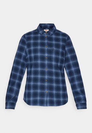 Chemise à manches longues, à boutons, en motif écossais bleu foncé. Présente un col, des boutons marron et un petit logo sur la poitrine gauche.